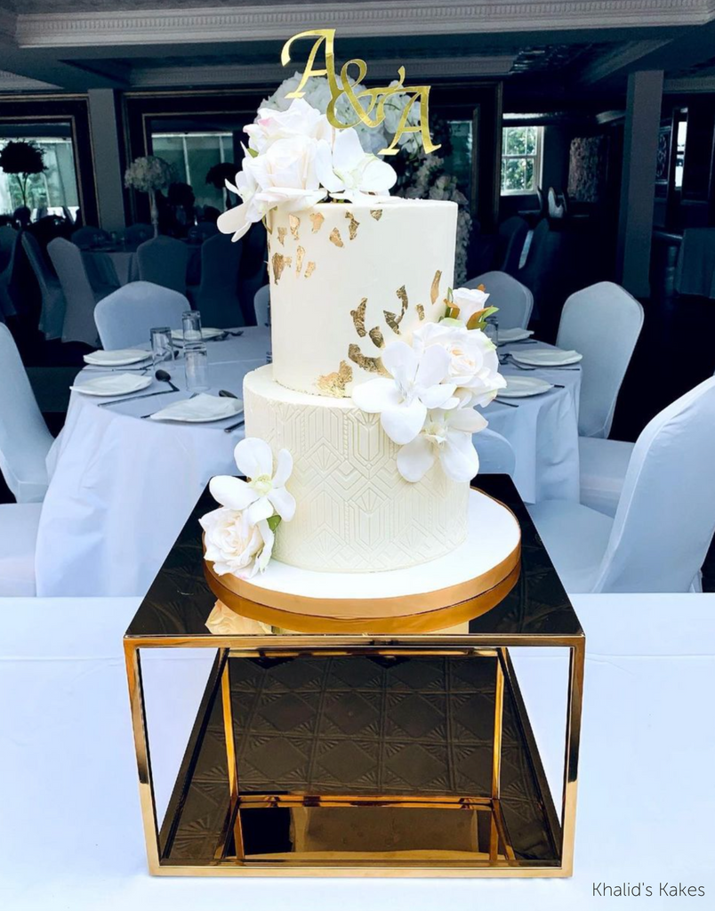 Gold Metallic Square Plinth holding up a white two tier cake covered in white flowers and gold decorations - Prop Options