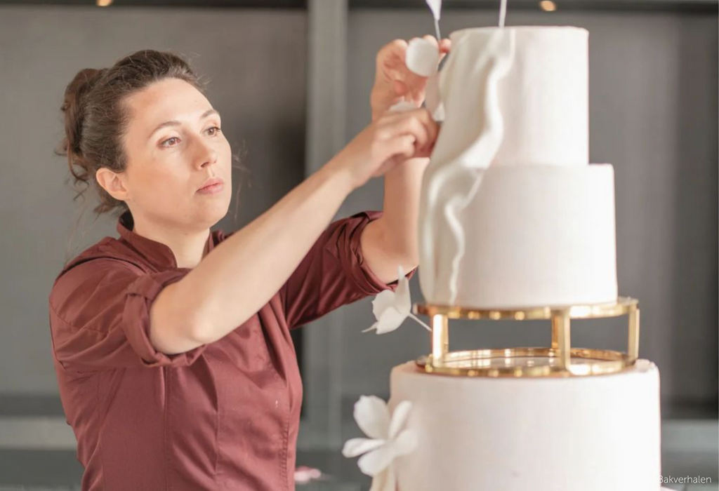 A baker adding white floral decorations to a white multitiered cake that uses a Gold 10" Round Metallic Cake Spacer in the middle of the tiers- Prop Options