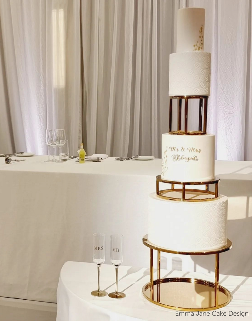 Gold Round Metallic Plinth holding up a tall multitiered wedding cake with subtle gold decorations and writing, the tiers are separated with gold spacers - Prop Options