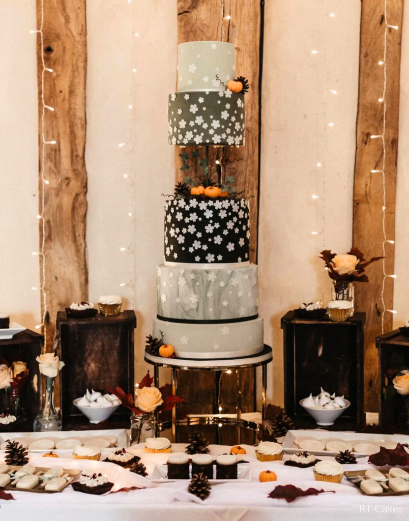 Gold Round Metallic Plinth holding up a tall Autumn themed cake with black and grey layers that are decorated with white flower emblems and mini orange pumpkins - Prop Options