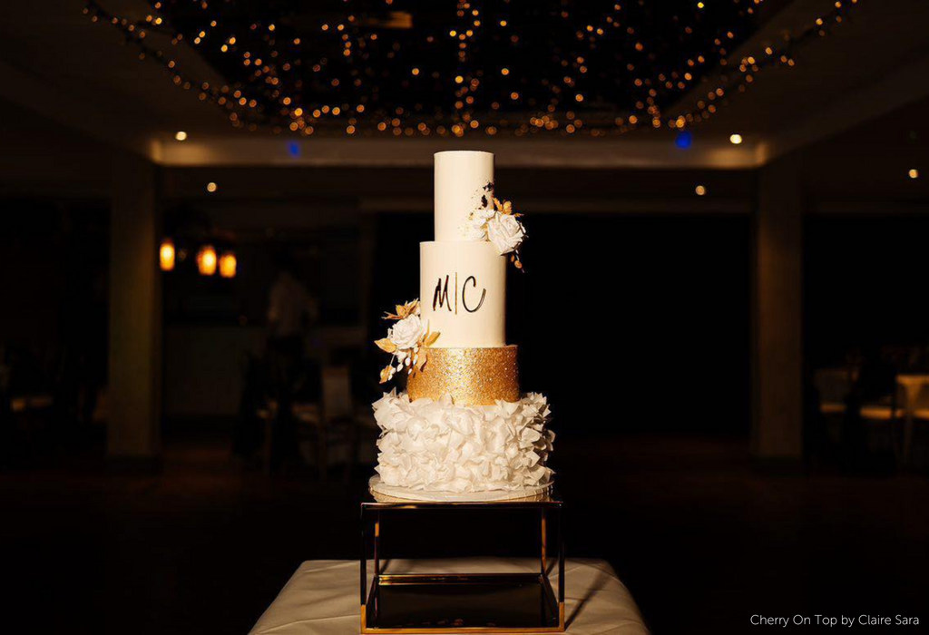 Gold Square Metallic Plinth holding up an elegant white cake with one gold tier, the cake has gold lettering and white and gold floral decorations on the side - Prop Options