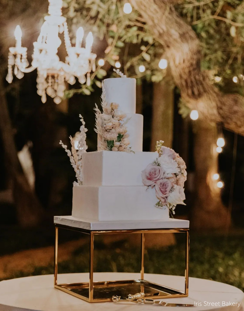 Gold Square Metallic Plinth holding up a white wedding cake with square bottom tiers and round top tiers, the cake is covered in floral decorations - Prop Options