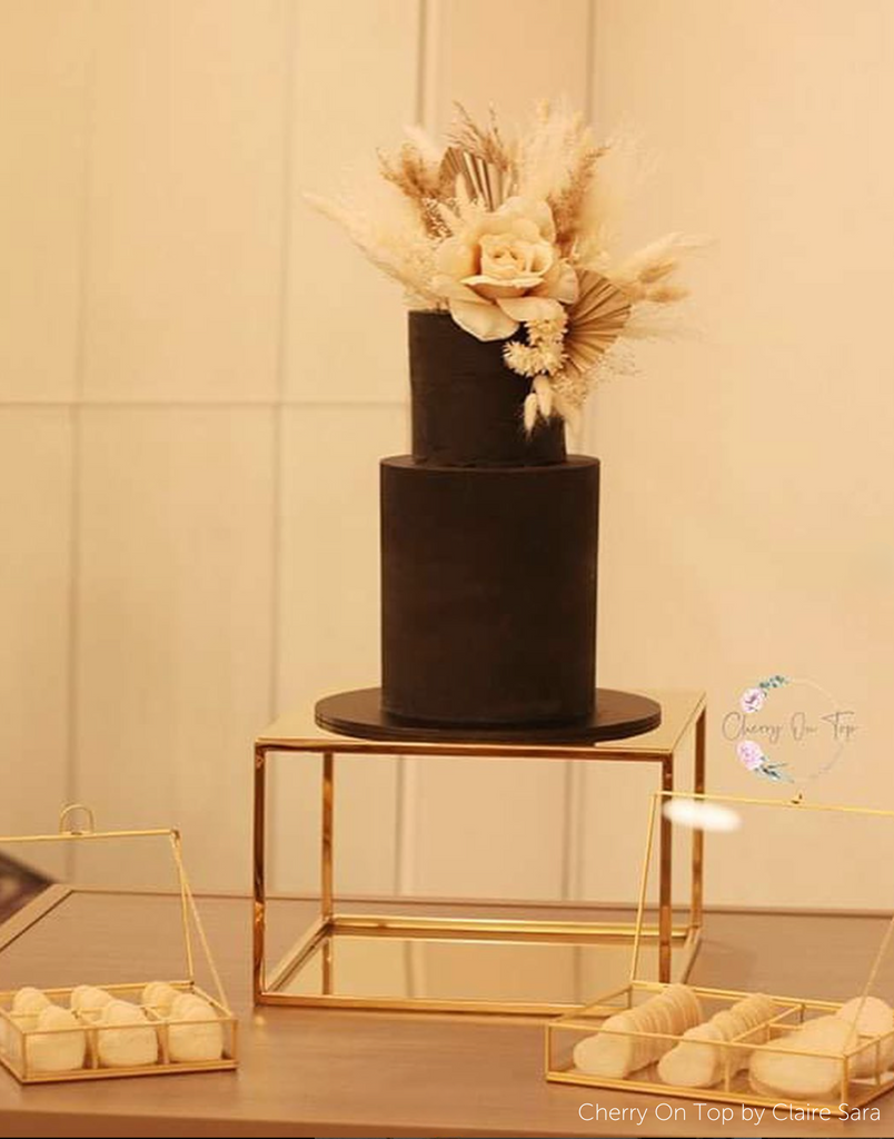 Gold Square Metallic Plinth holding up a black two tier cake with cream coloured floral decorations on the top - Prop Options