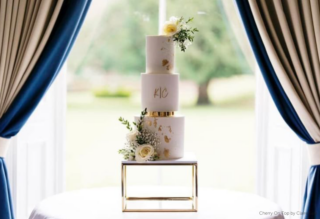 Gold Square Metallic Plinth holding up a white three layer cake with gold decorations and lettering, with white roses on the top and base - Prop Options
