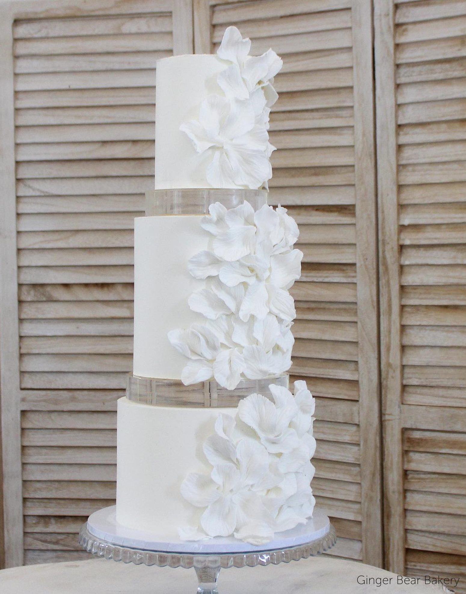 A plain white three tier cake with matching white floral decorations down the side, each layer is separated with a 30mm Round Acrylic Cake Separator - Prop Options