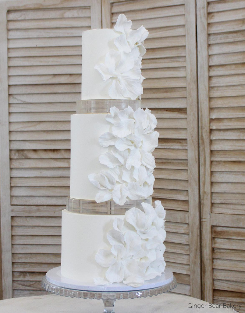 A plain white three tier cake with matching white floral decorations down the side, each layer is separated with a 30mm Round Acrylic Cake Separator - Prop Options