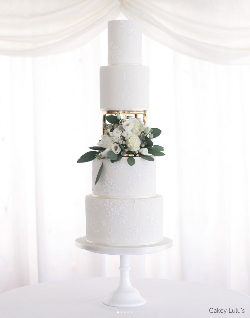 Plain white four tier cake with subtle floral designs in the icing separated in the middle by a 6" Round Metallic Cake Spacer covered in white roses - Prop Options