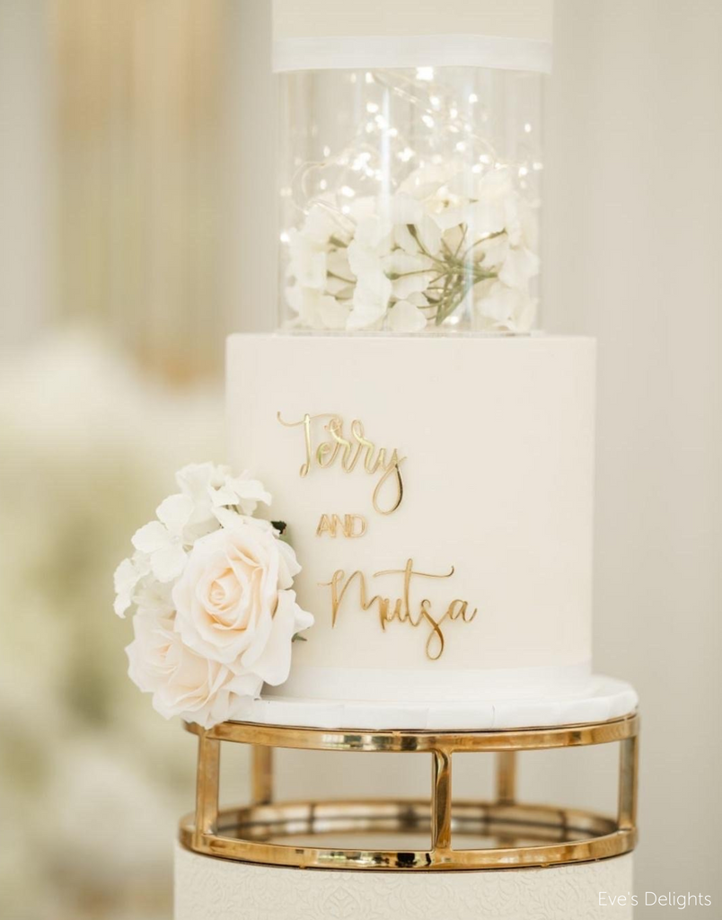 White wedding cake that has names embossed in gold with acryclic tier filled with flowers held up by 10" Round Metallic Cake Spacer - Prop Options