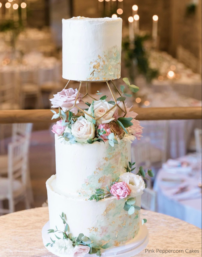 A white cake with soft pastel decorations and flowers along the side, the top two layers are separated using a Geometric Pentagon Spacer - Prop Options