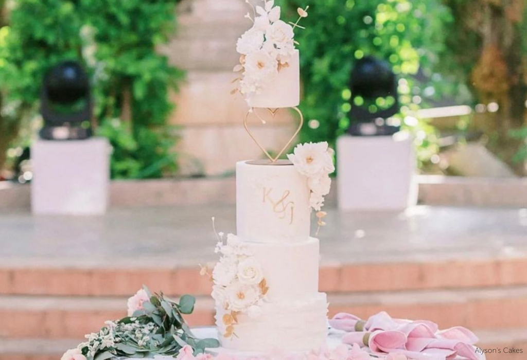 A white four layer cake with white flower decorations and the initials K & J in gold lettering on the front, the top two layers are separated by a Heart Tier Separator - Prop Options