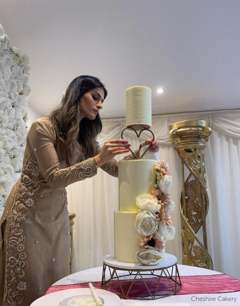 A plain cream coloured three tier cake with cream and peach flowers along the side, a woman is adjusting a Heart Tier Separator between the top two layers - Prop Options