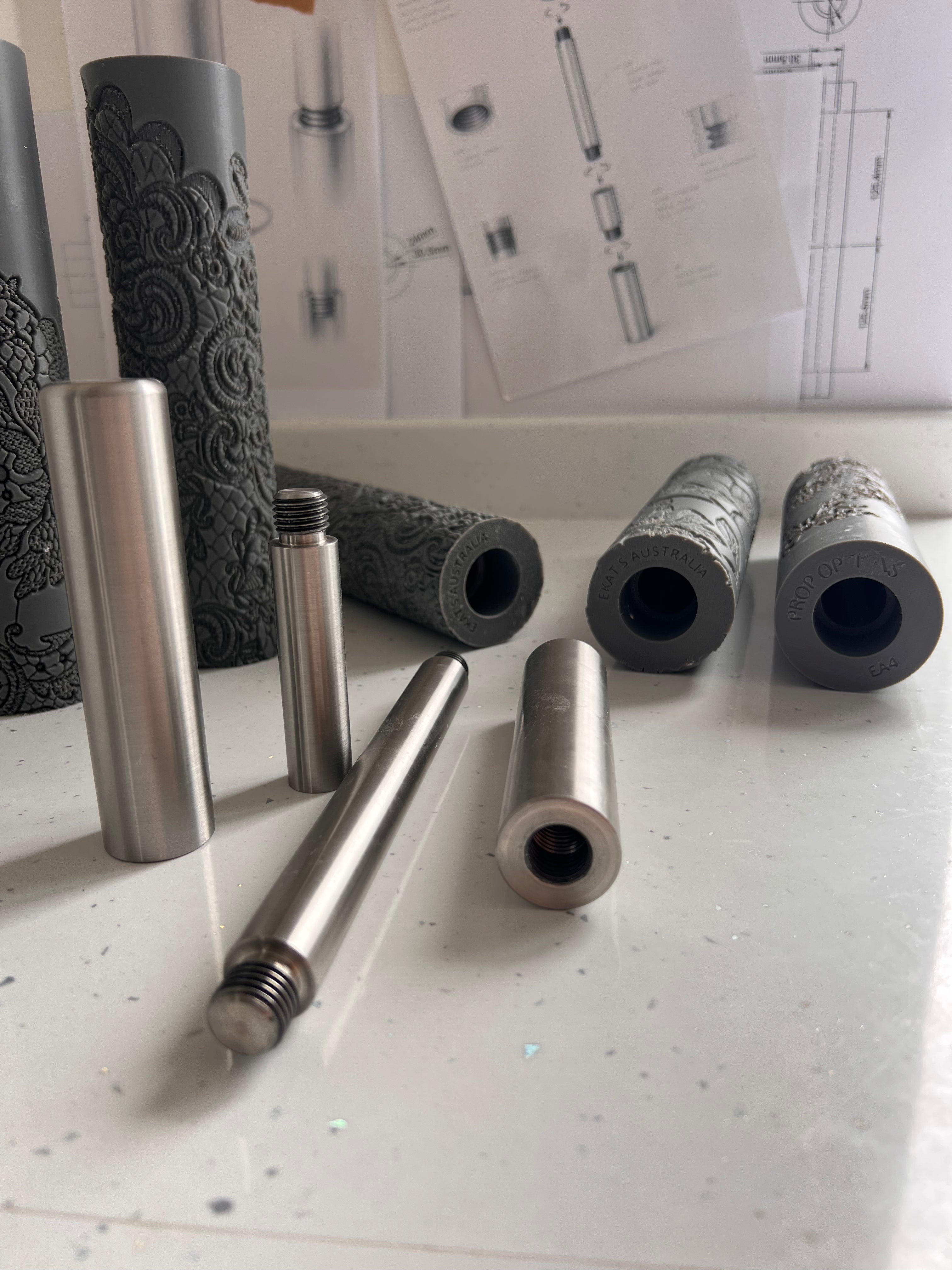 Metal and foam cylindrical objects on a white surface with technical drawings in the background.