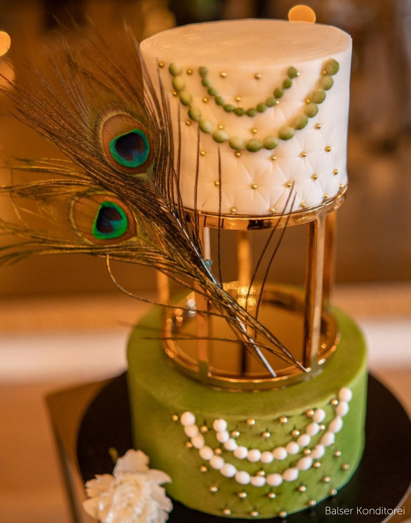 A two tier cake with a green bottom tier covered in white decorations and a white top tier covered in green decorations, the tiers are separated using a 6" Round Metallic Cake Spacer with a peacock feather attached - Prop Options