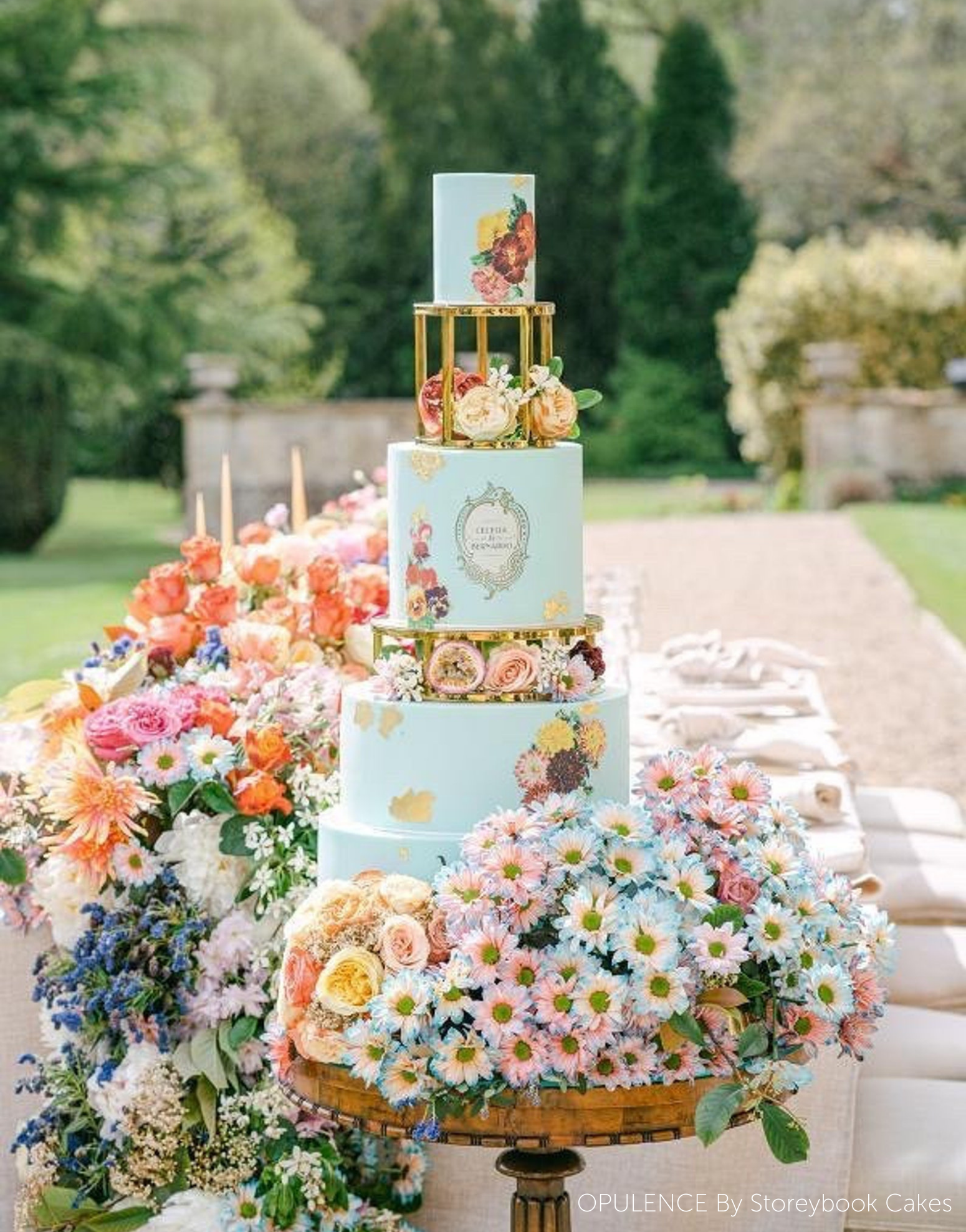 A light blue multitiered cake with pastel coloured flowers, the tiers are separated using 6" & 10" Round Metallic Cake Spacers - Prop Options