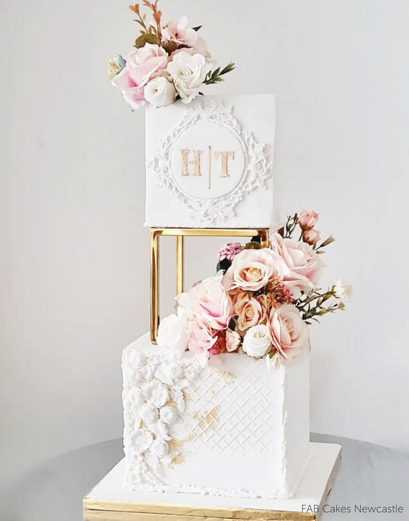 A square white two layer cake with gold lettering and pink flowers being separated by a Square Metallic Cake Spacer - Prop Options