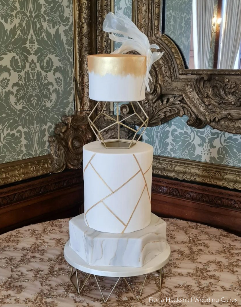 Geometric Cake Stand holding a three layer cake, the bottom layer is marbled white, grey, and goold, the middle layer has gold accents, and the top layer is gold and white, between the top layers is a geometric spacer - Prop Options