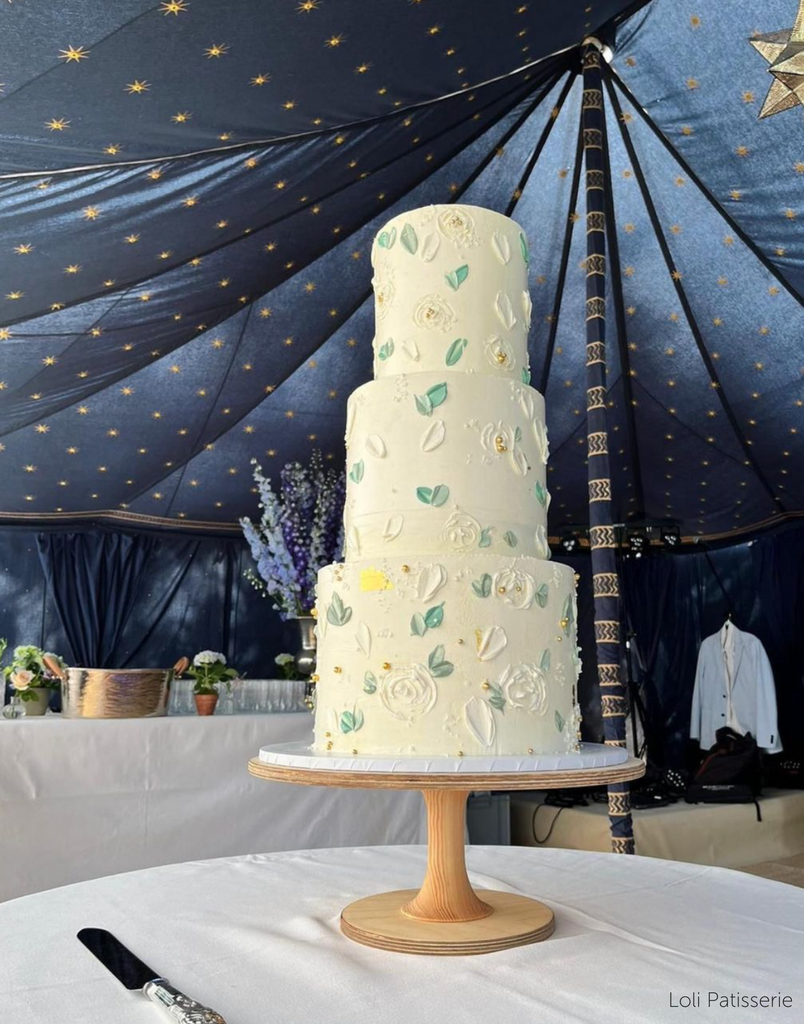 White cake with embedded green leaves and white florals stood on The Hourglass Scandinavian Birch Cake Stand - Prop Options
