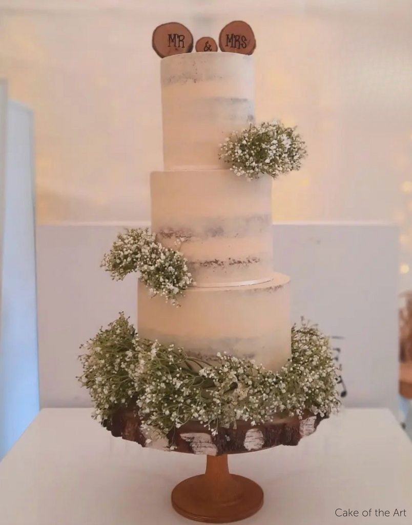 A rustic white cake with green leaves stood on The Hourglass Scandinavian Birch Cake Stand - Prop Options