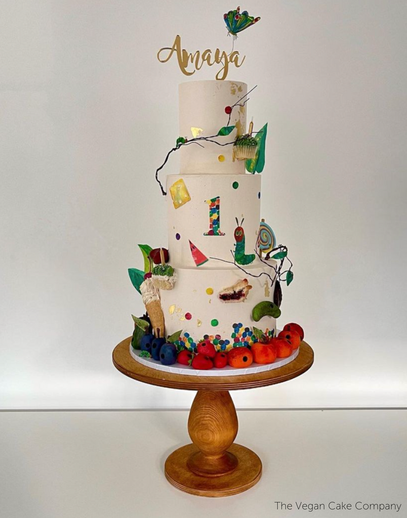A white cake covered in fruit and caterpillar decals stood on The Teardrop Scandinavian Birch Cake Stand - Prop Options