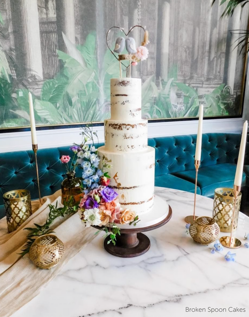 A rustic white cake with colourful flowers along the bottom and a heart topper with two birds stood on The Teardrop Scandinavian Birch Cake Stand - Prop Options