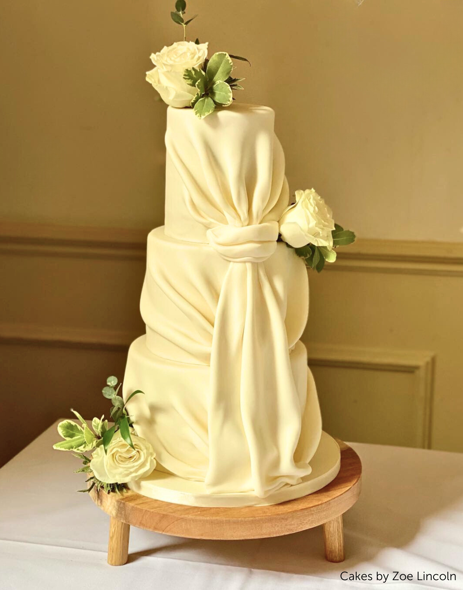 A tall cake covered in cream coloured fondant and white roses on top of a Solid Oak Tripod Cake Stand - Prop Options