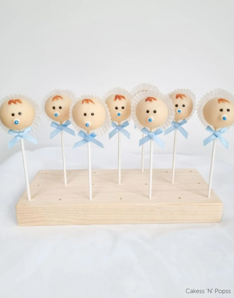 Cake pops decorated to look like baby boys with white bonnets and blue ribbons being held in an English Pine Wooden Cake Pop Stand - Prop Options